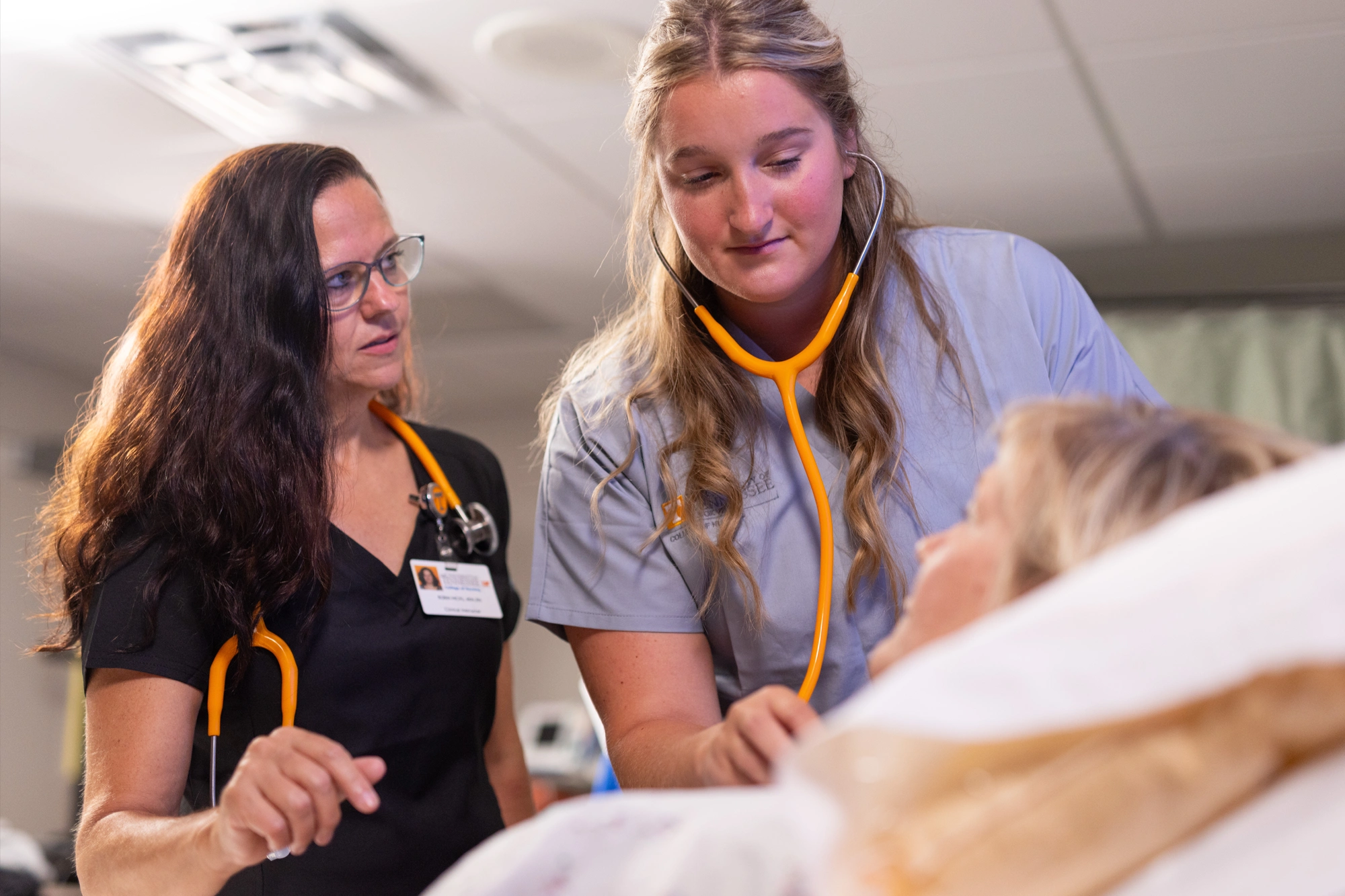 One woman takes a patient's heart rate with a stethoscope while another woman watches her